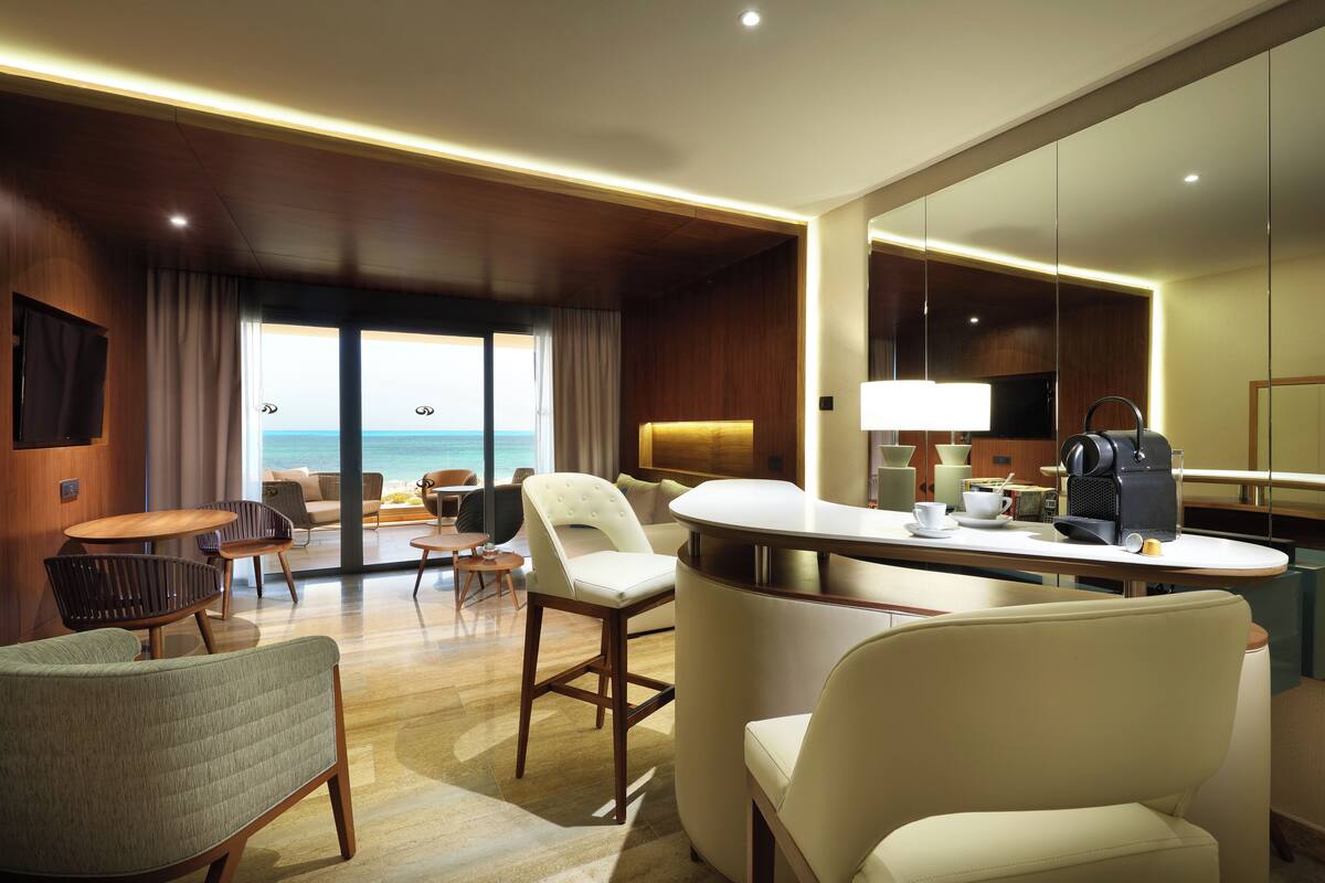 A modern hotel room with a large window offering a view of the ocean, a sitting area with chairs and a table, and a kitchenette with a coffee machine.