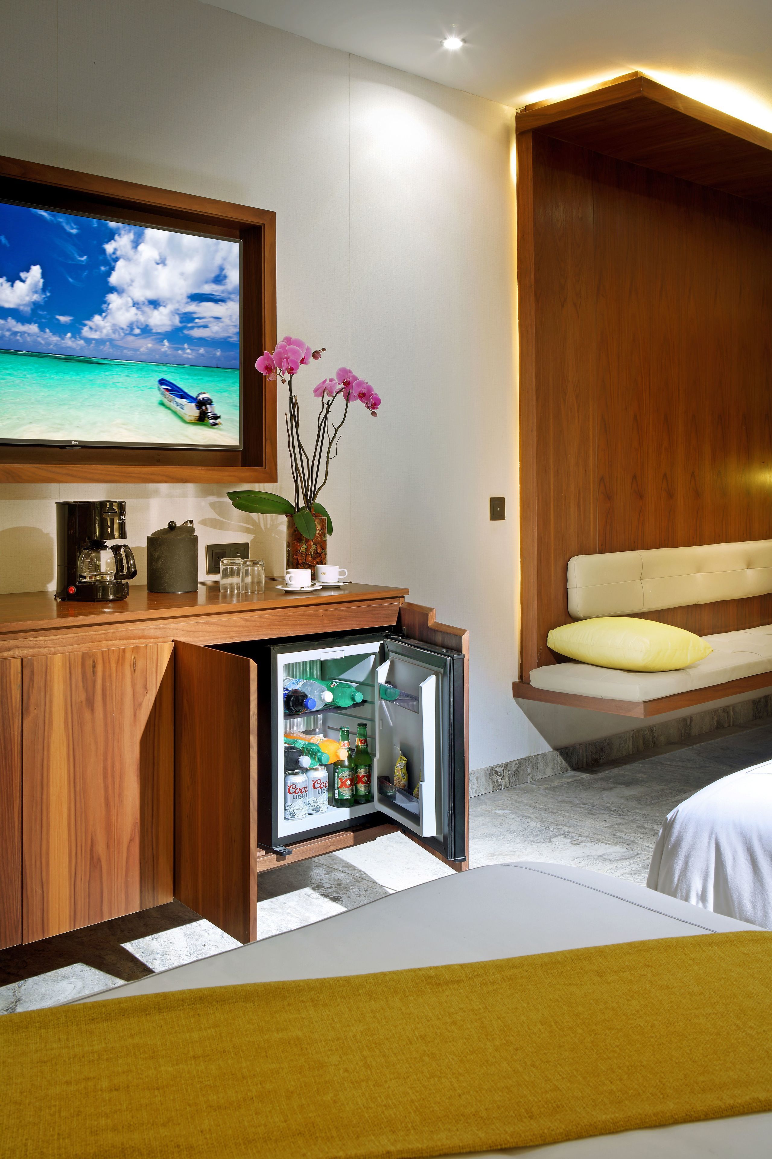 Habitación de hotel con escritorio de madera, refrigerador pequeño, televisor mostrando una escena de playa, cama con almohada amarilla y banco de madera.