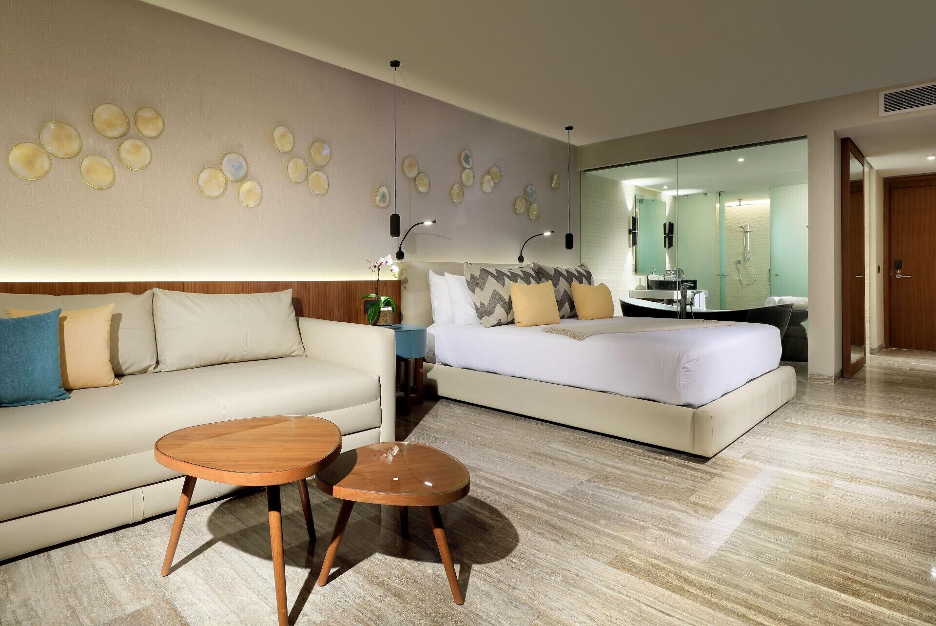 A hotel room with a bed, a sofa, a small table, a mirror, and a wall with circular patterns.