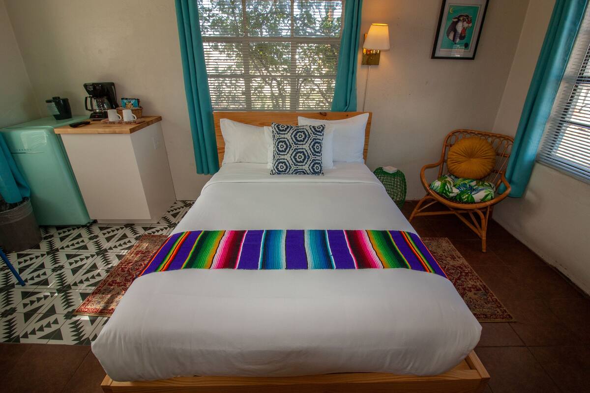Un lit bien fait, recouvert d’une couverture rayée aux couleurs vives, une petite table de chevet avec une machine à café et une chaise avec un coussin.