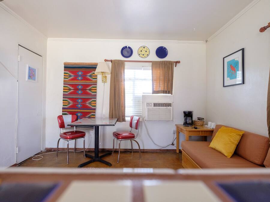 A compact living space with a dining table, two chairs, a sofa, a window with curtains, and a wall-mounted air conditioner.