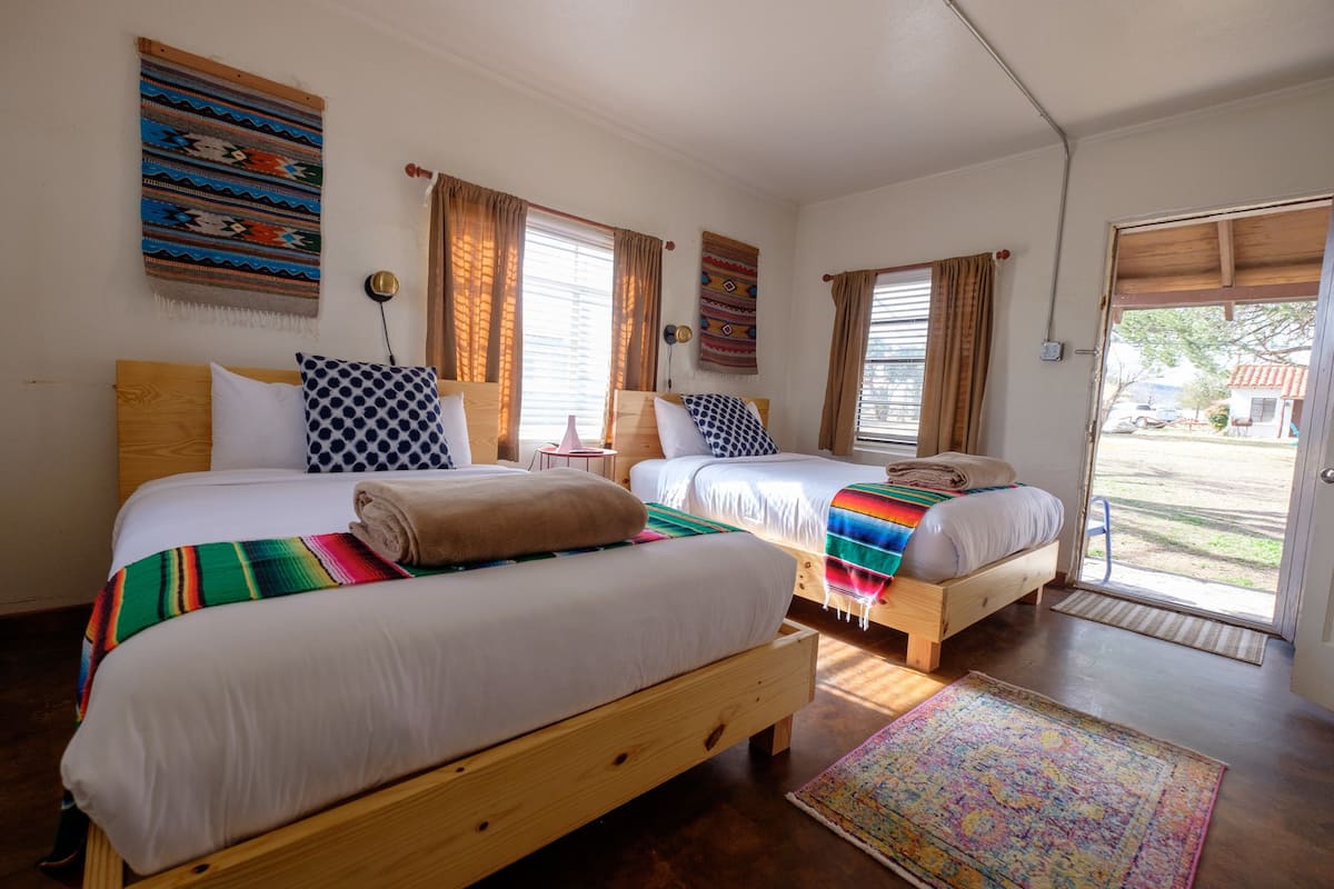 Une pièce avec deux lits en bois, chacun doté d’un matelas blanc et d’une couverture colorée. On y trouve deux fenêtres avec des rideaux, une lampe fixée au mur et une porte donnant sur l’extérieur.