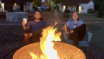 Two people sitting by a campfire in an outdoor setting with a stone structure and string lights in the background.