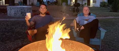 Two people sitting by a campfire in an outdoor setting with a stone structure and string lights in the background.