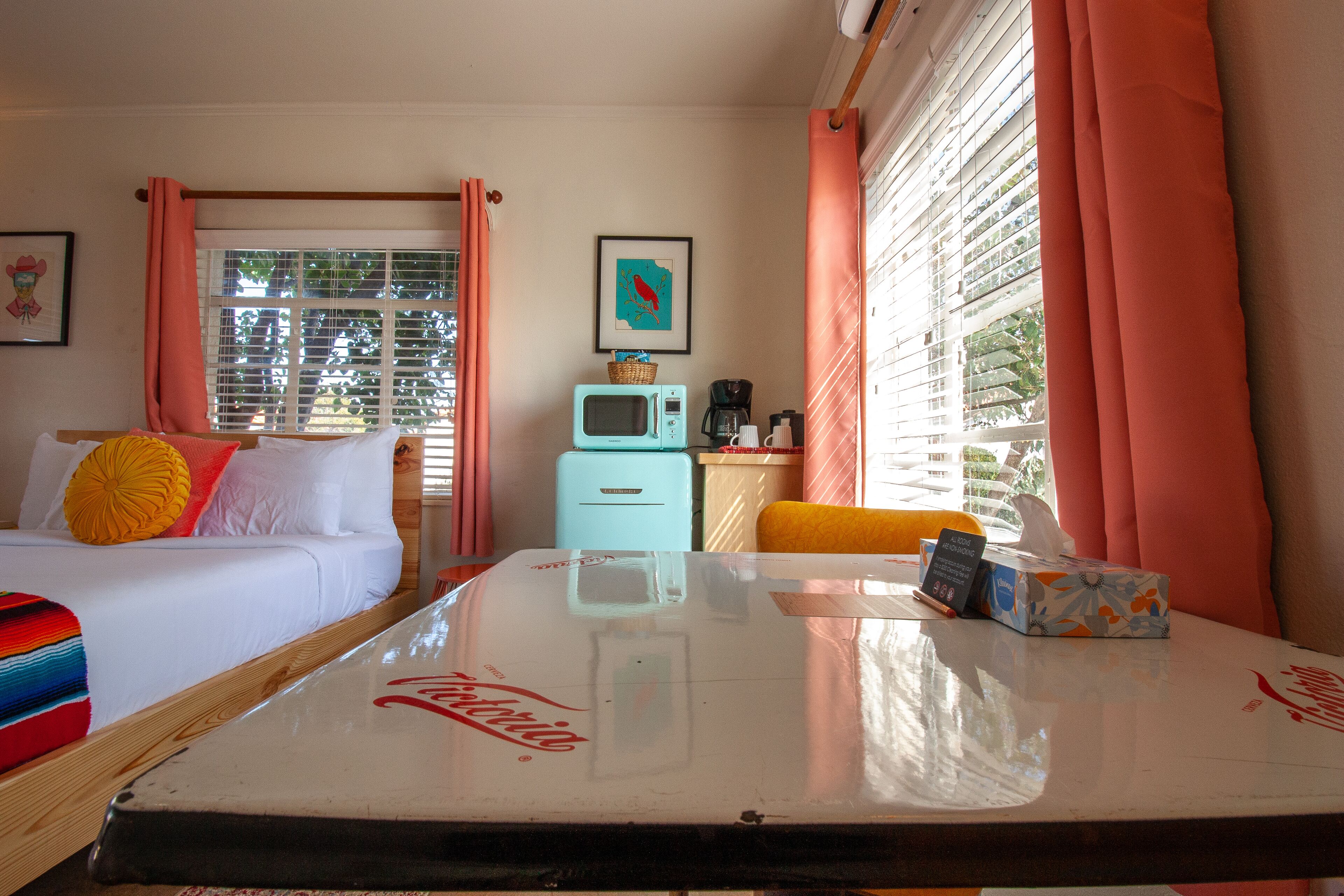 A hotel room with a bed, a small kitchenette, a window with curtains, and a table with a Coca-Cola logo.