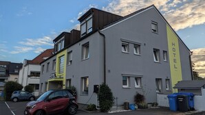 Et moderne hotel med en grå facade, en gul accentvæg og et skilt, der angiver, at det er et hotel. Der er biler parkeret foran, og en rød bil oplades.