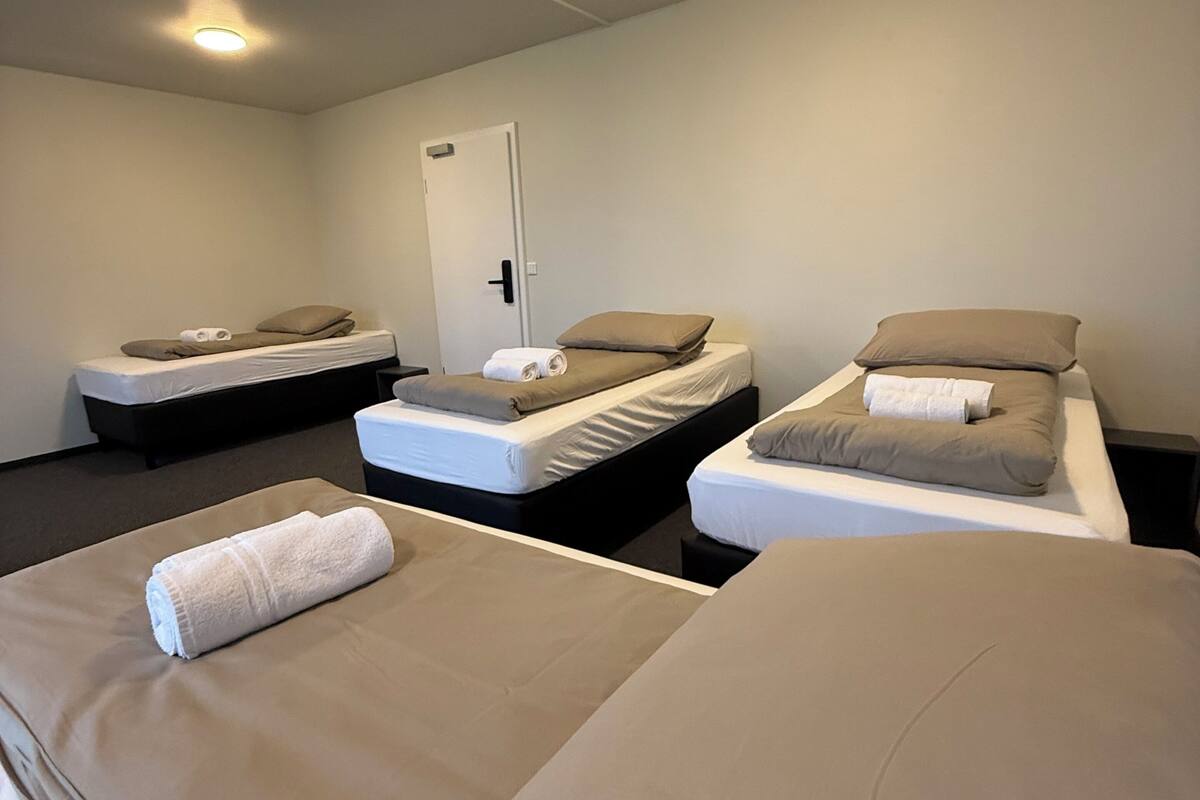 A hotel room with three single beds, each with a pillow and a rolled towel. There is a door in the background and a ceiling light.