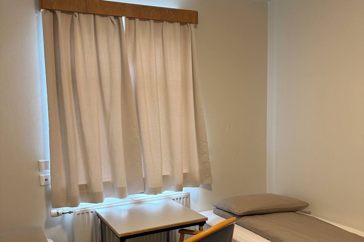 A small, compact room with a single bed, a chair, a table, and a radiator.