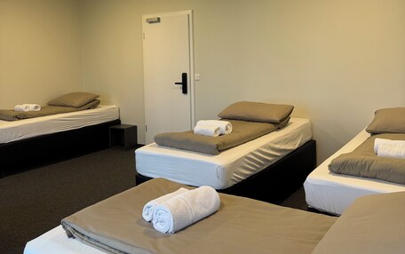 A hotel room with three single beds, each with a pillow and a rolled towel. There is a door with a handle and a light fixture on the ceiling.