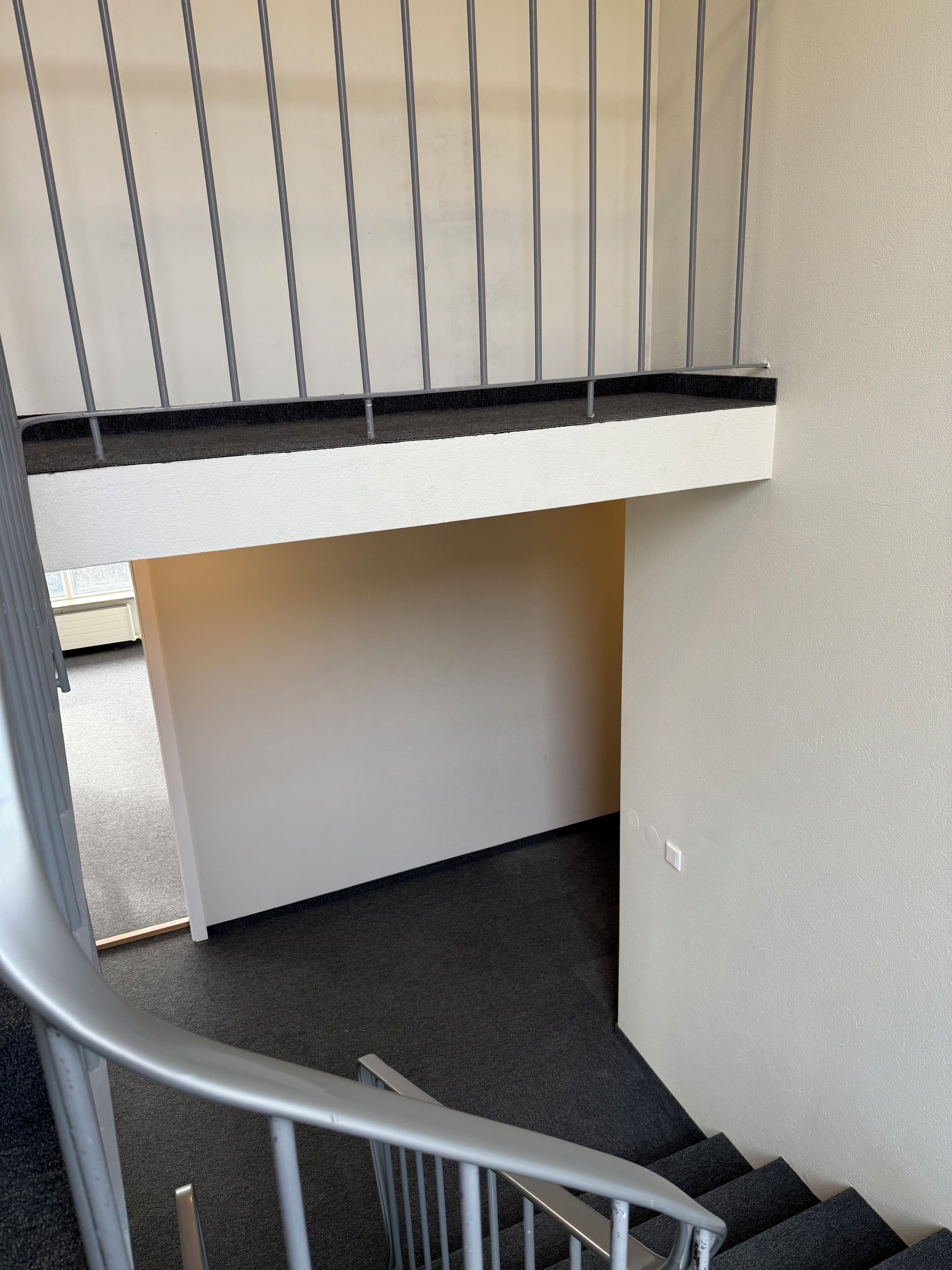 Una escalera con barandilla metálica, peldaños alfombrados y una pared blanca con interruptor de luz.