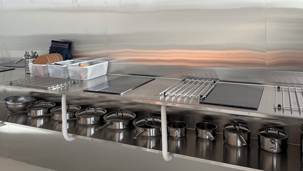 A stainless steel kitchen with a central island, overhead ventilation, and a row of pots below.
