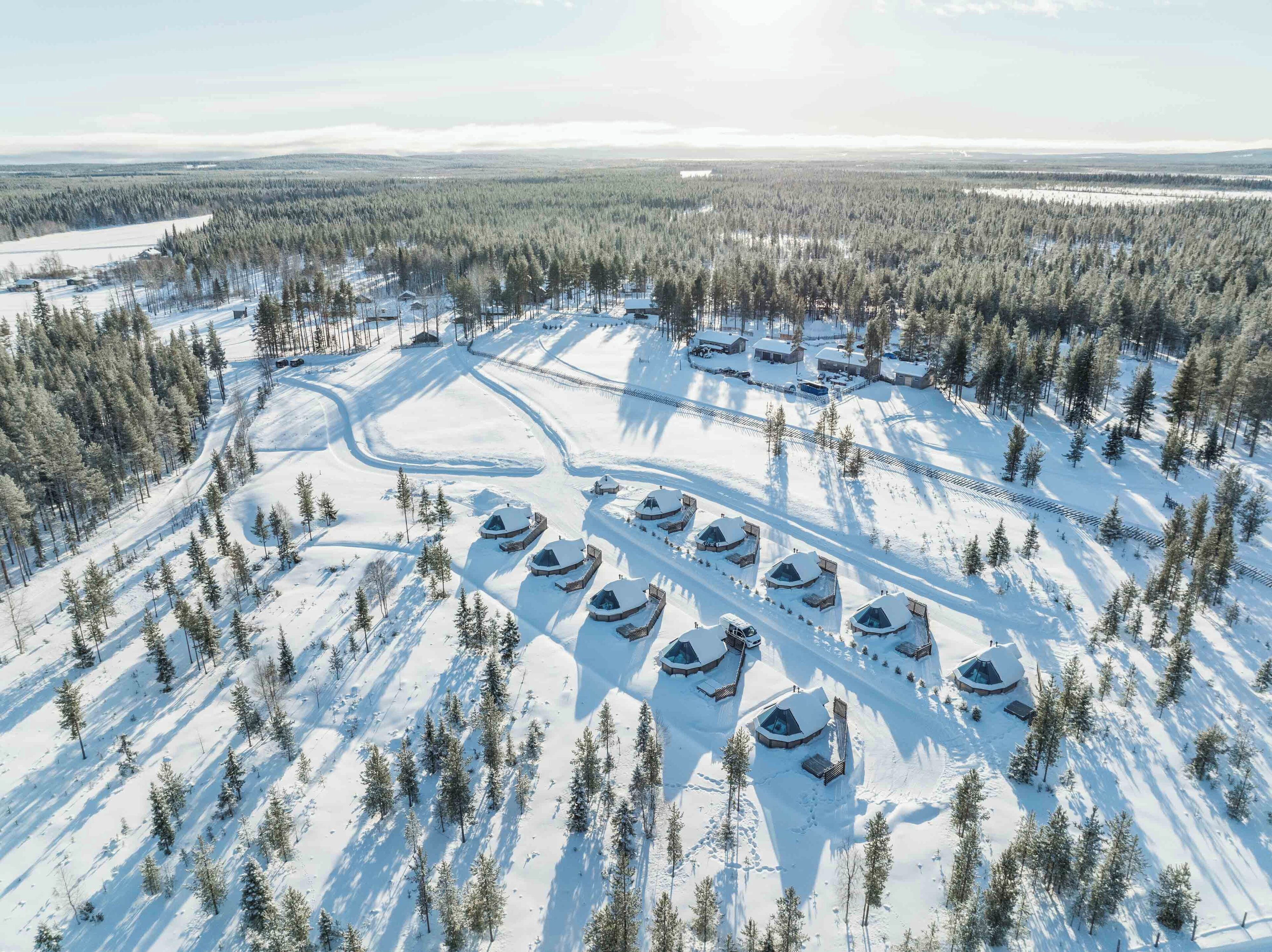 An aerial view of a snow-covered area with several circular cabins arranged in a semi-circular pattern, surrounded by dense forest.