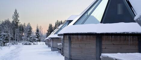 A row of wooden cabins with snow-covered roofs and icicles, surrounded by a snowy forest.