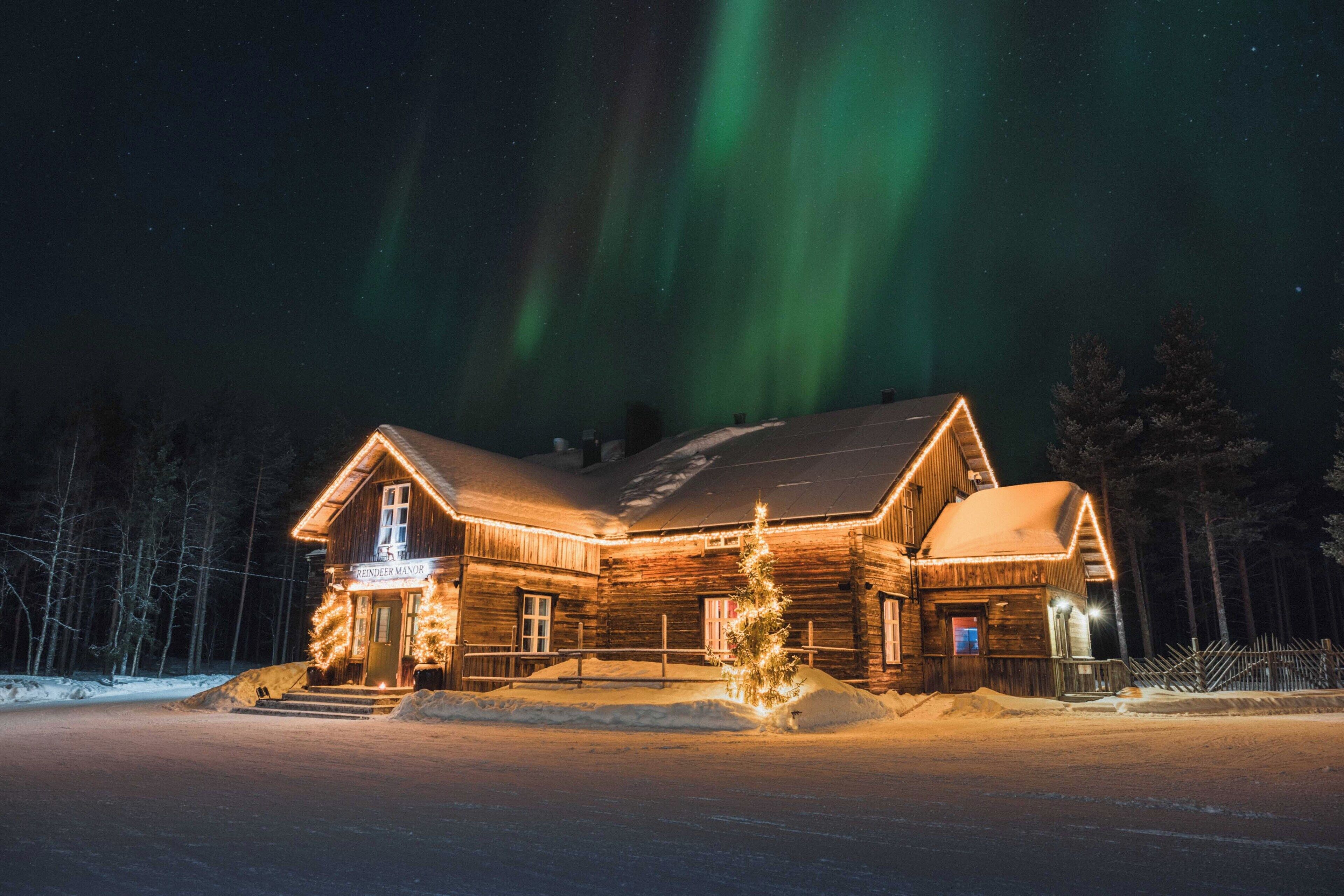 A log cabin with a lit entrance and a Christmas tree, surrounded by snow and trees.