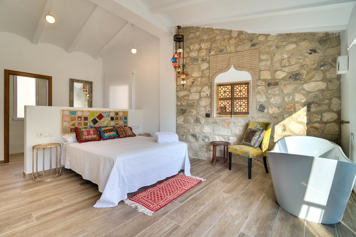 A bedroom with a stone wall, a bed with colorful pillows, a red rug, a yellow chair, and a white bathtub.