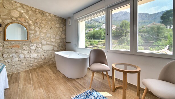 Un baño con pared de piedra, una bañera y vistas a montañas a través de amplios ventanales.