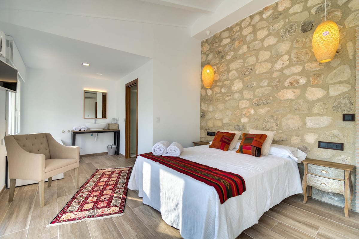 A bedroom with a large bed, a sitting area with a chair, a stone wall, and a bathroom with a sink and mirror.