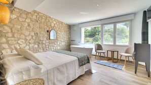 Un dormitorio con pared de piedra, una cama grande con un edredón azul y blanco, una bañera y vistas a la vegetación a través de las ventanas.
