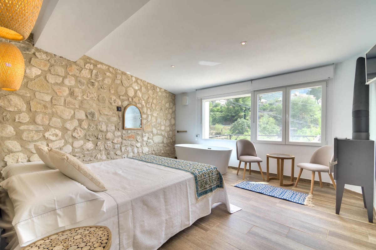A bedroom with a stone wall, a large bed with a blue and white throw, a bathtub, and a view of greenery through the windows.