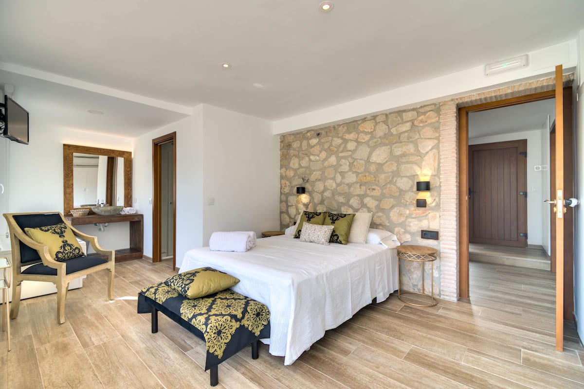 A bedroom with a large bed, a chair, a small table, and a stone wall.