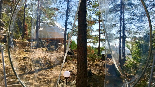 A transparent spherical structure with a wooden platform and a spherical light fixture, surrounded by trees and a rocky terrain. - FinnDome- Glamping (Pori)