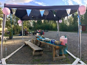 Un espace de restauration en plein air au décor rustique, avec une table et des bancs en bois, entouré d’une tente décorée de ballons roses.