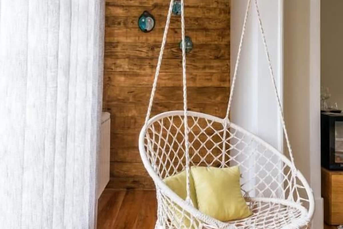 A white macrame hanging chair with green cushions in a room with wooden walls and a white curtain.