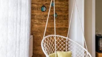 Una silla colgante de macramé blanco con cojines verdes en una habitación con paredes de madera y una cortina blanca.