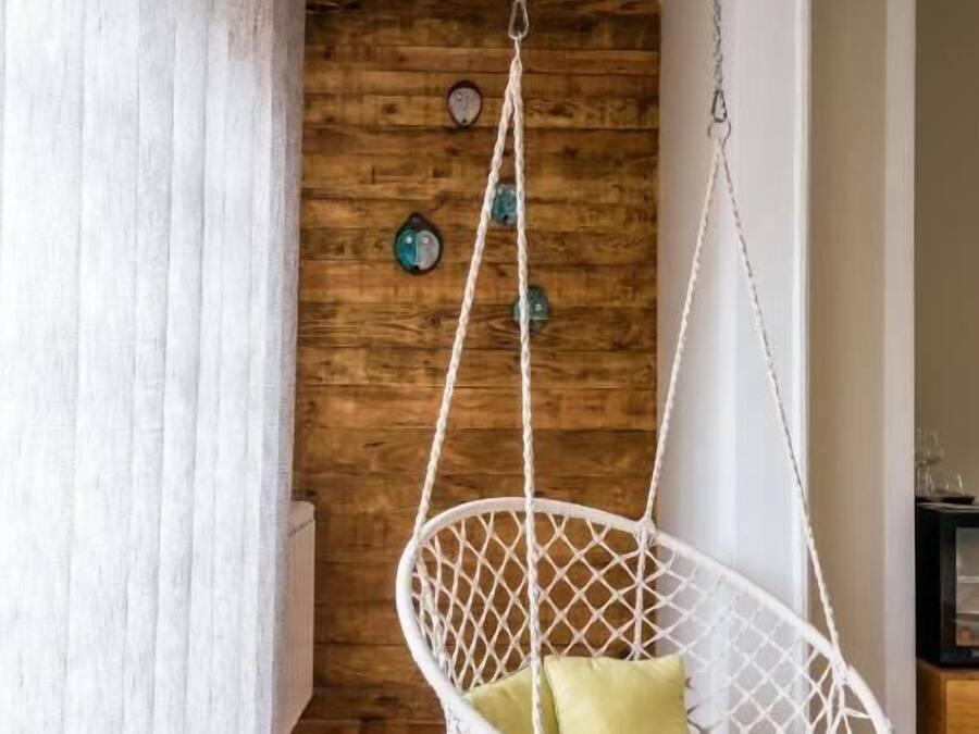 Una silla colgante de macramé blanco con cojines verdes en una habitación con paredes de madera y una cortina blanca.