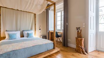 Une chambre à coucher avec un lit à baldaquin, un plancher en bois, un bureau avec une chaise et un tabouret en bois agrémenté d’un objet décoratif.