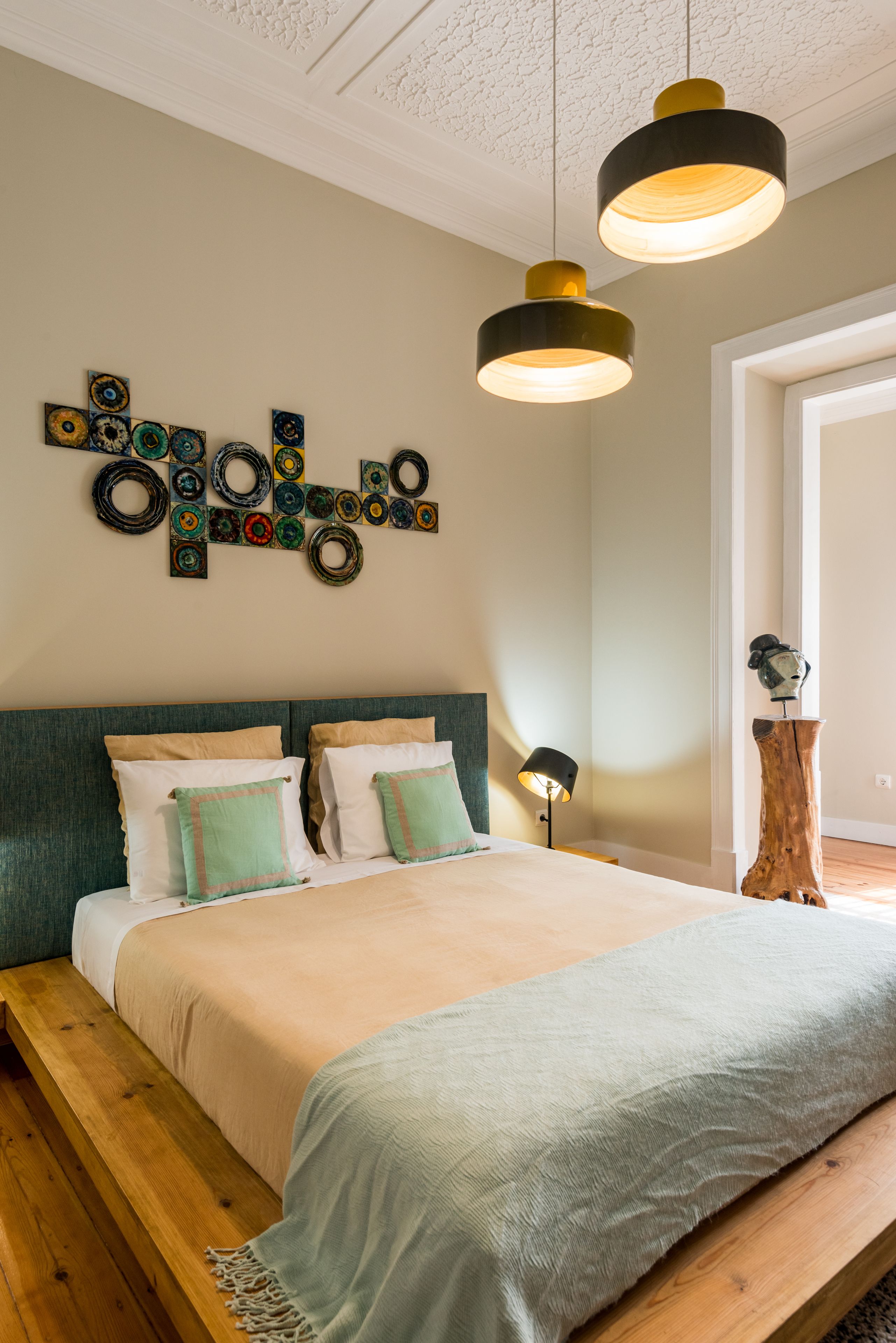 Quarto com cama, banco de madeira, obras de arte na parede e luminárias pendentes.
