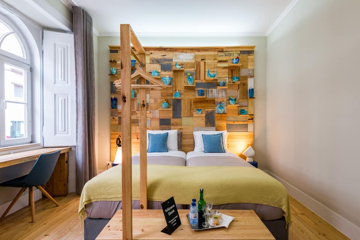 A hotel room with a wooden wall display, a bed with a green blanket, a desk with a chair, and a bedside table with a welcome sign and drinks.