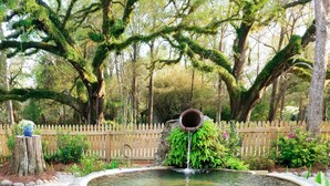 Een ronde stenen fontein met een centraal pijpje, omringd door een houten hek en weelderige begroeiing.