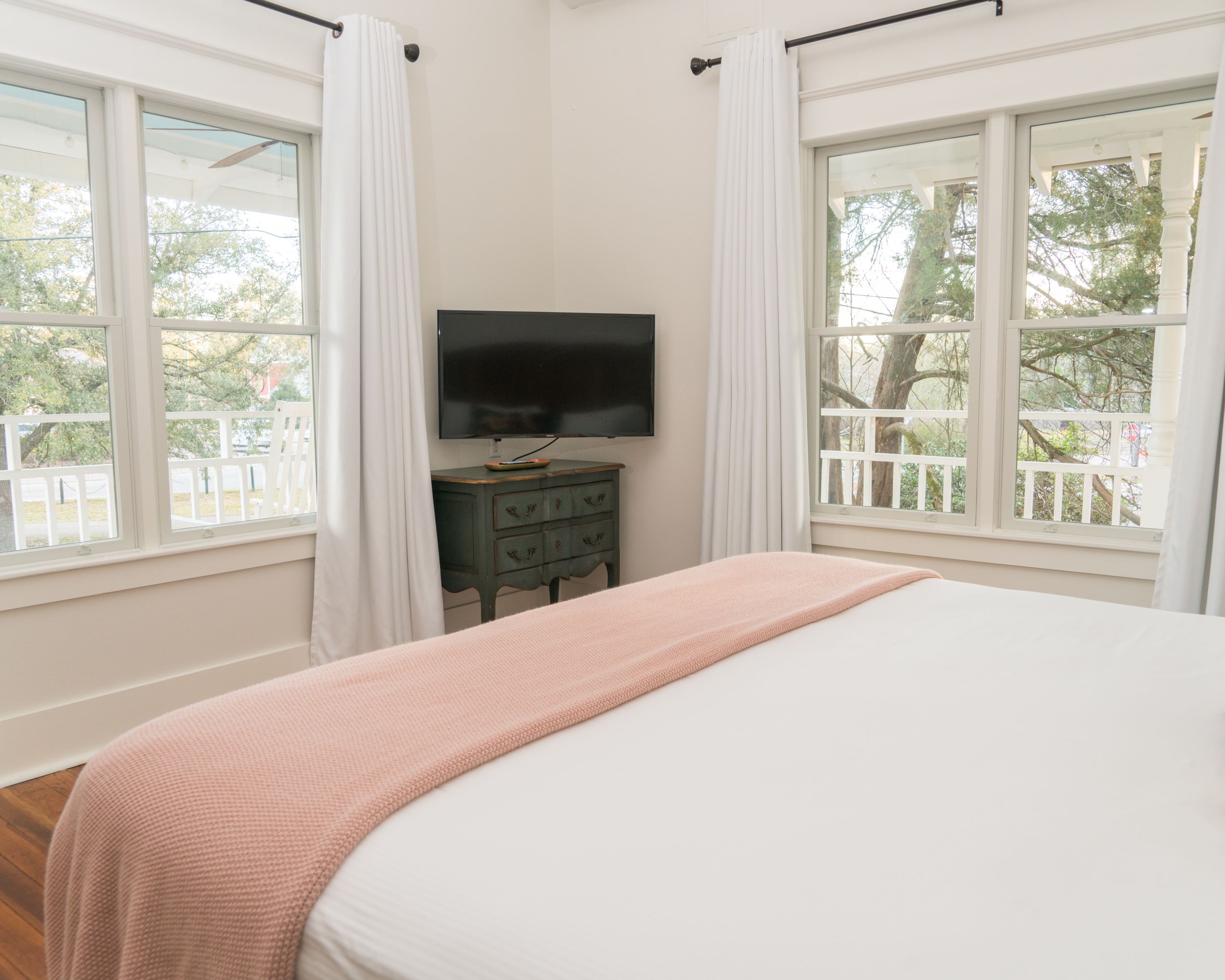 A bedroom with a bed, a dresser, a TV, and large windows with curtains.