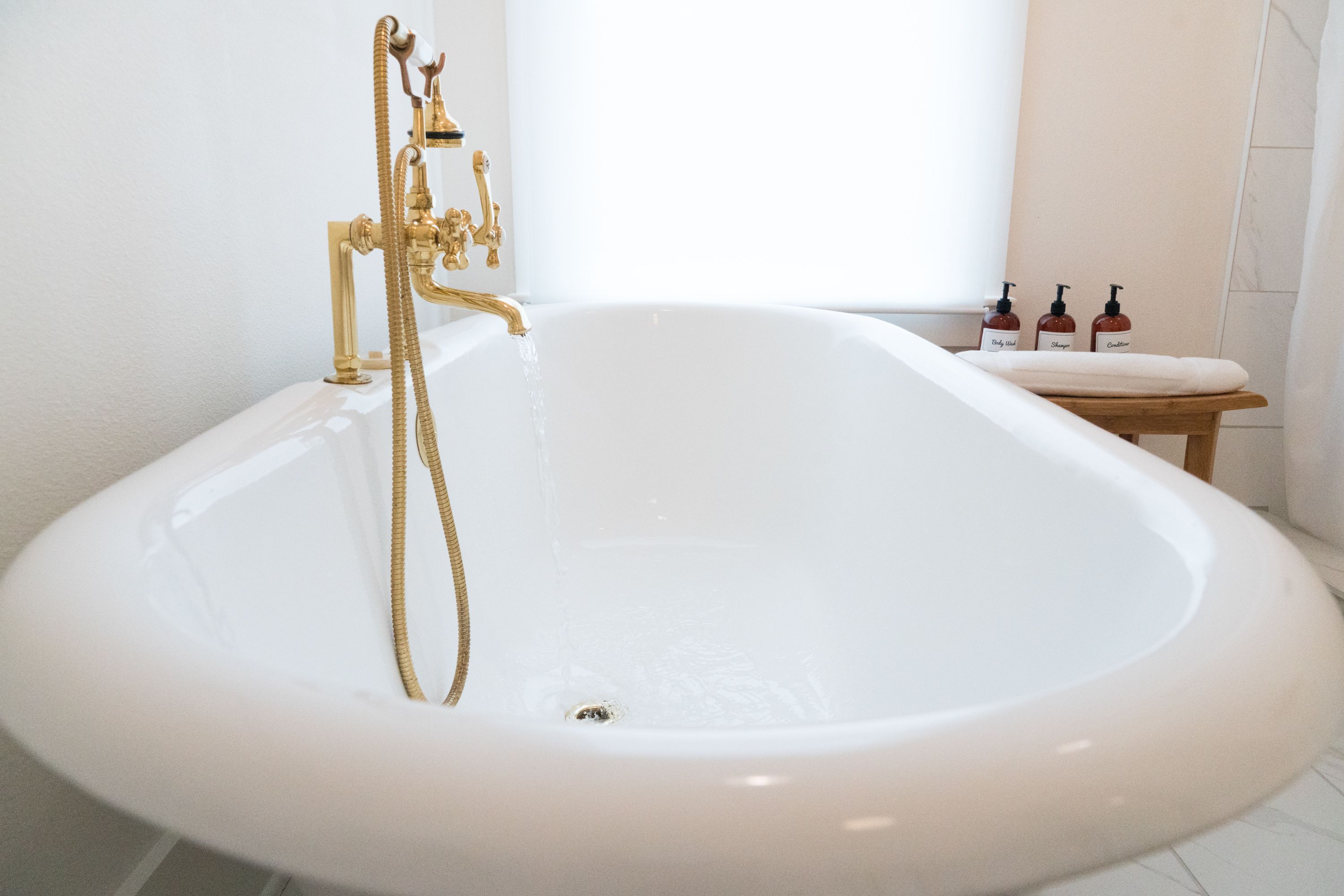 A large, oval-shaped bathtub with a gold faucet and hose, a wooden stool, and three bottles of shampoo on the side.