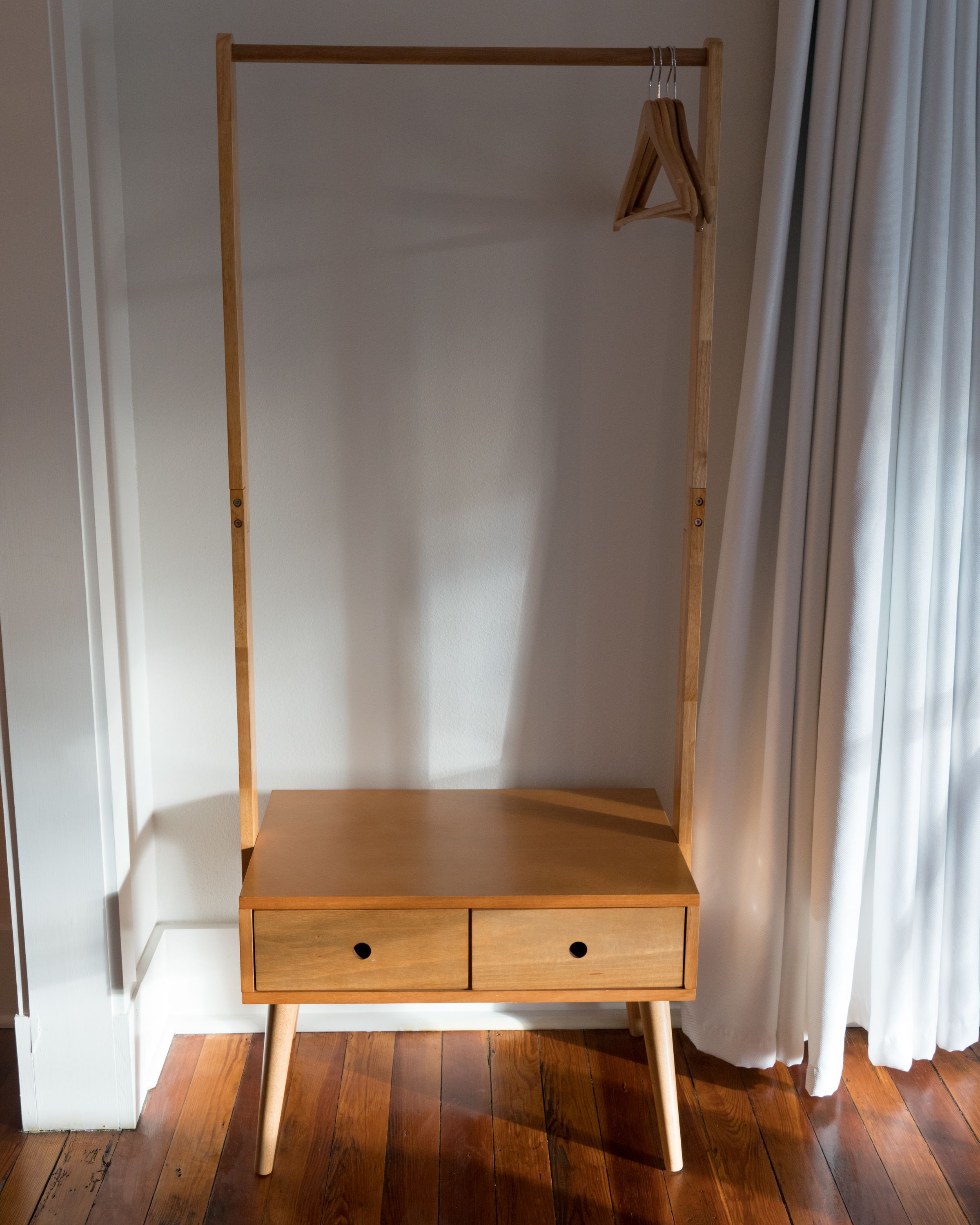 A wooden wardrobe with two drawers and a hanging space, positioned against a white wall with a wooden floor.