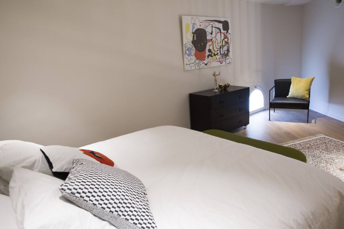 A bedroom with a large bed, a black dresser, a chair with a yellow cushion, and a wall-mounted abstract painting.