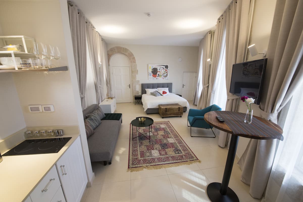 A hotel room with a bed, a sofa, a small table, a TV, a kitchen area with a stove, and a dining table with glasses.