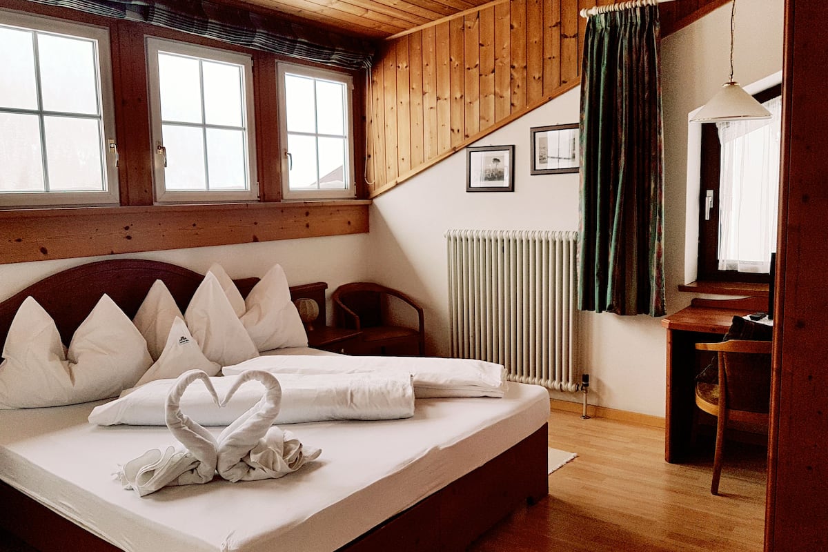 A bedroom with a wooden ceiling, a bed with white bedding, a heart-shaped towel arrangement, a desk, and a chair.