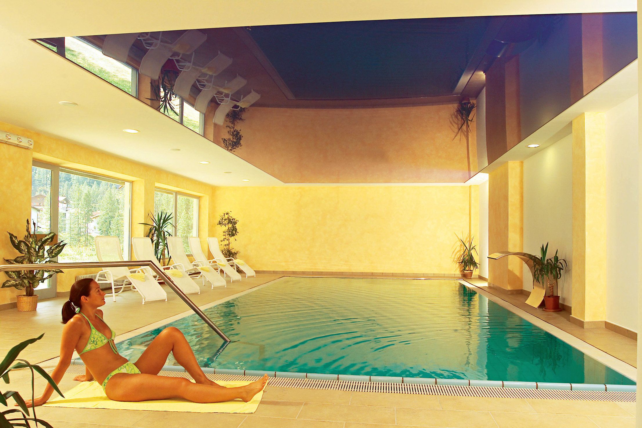 Ein Hallenbad mit einer Frau, die auf einer Matte sitzt. Um den Pool herum befinden sich Liegestühle und Kübelpflanzen.