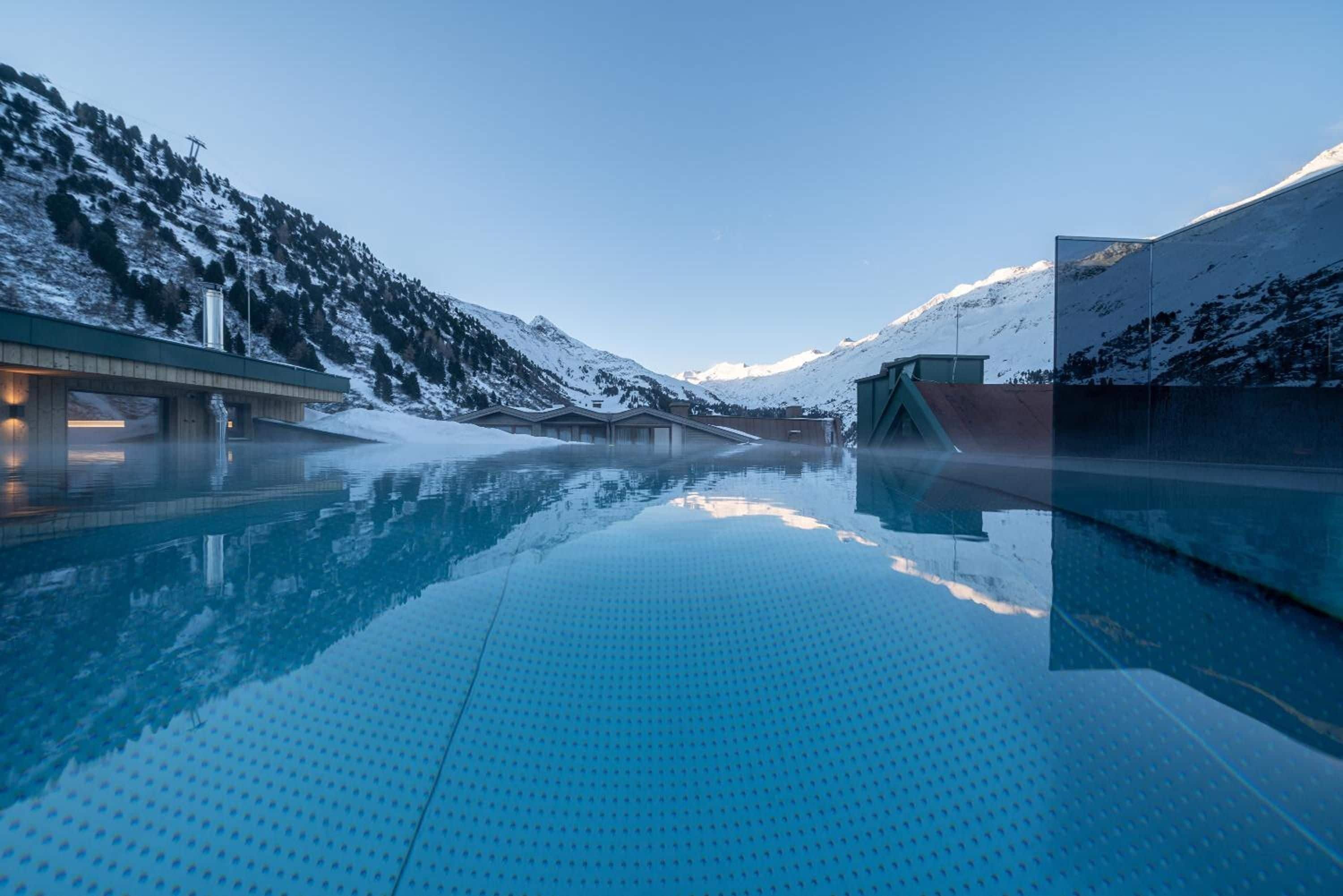 A clear pool with a mountain backdrop, a bridge, and a building with a sloped roof.