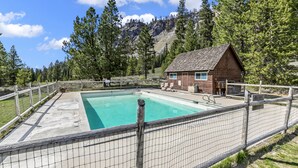 Una cabaña de madera con piscina, rodeada por una valla de madera y árboles, con montañas al fondo.