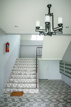 A staircase with patterned tiles, a fire extinguisher, a chandelier, and a fire alarm panel. - Lullaby Residence by Smarnbook (Phayakkhaphum Phisai)