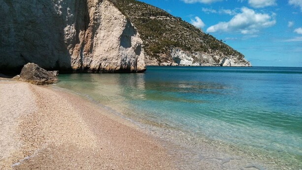 Una playa con agua turquesa cristalina, orilla de guijarros y un acantilado grande.