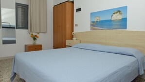Un dormitorio con cama, un armario de madera, una mesita de noche con un florero, un cuadro de acantilados y una ventana con cortinas.