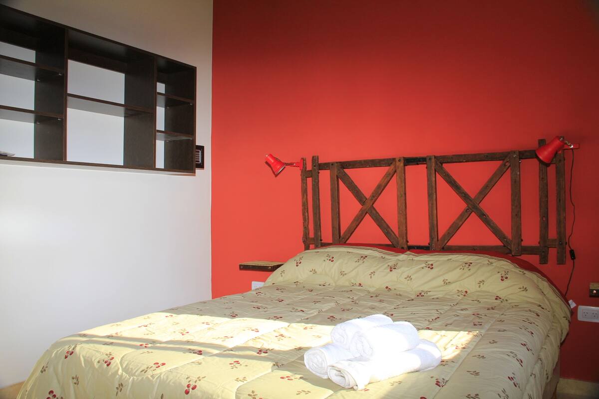 A bed with a floral bedspread, a wooden headboard, and a red wall.