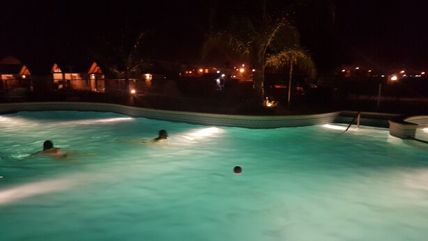 Una piscina con tres personas nadando, rodeada de edificios iluminados por la noche.