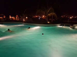 Une piscine où se trouvent trois personnes, entourée de bâtiments dont les lumières sont allumées la nuit.