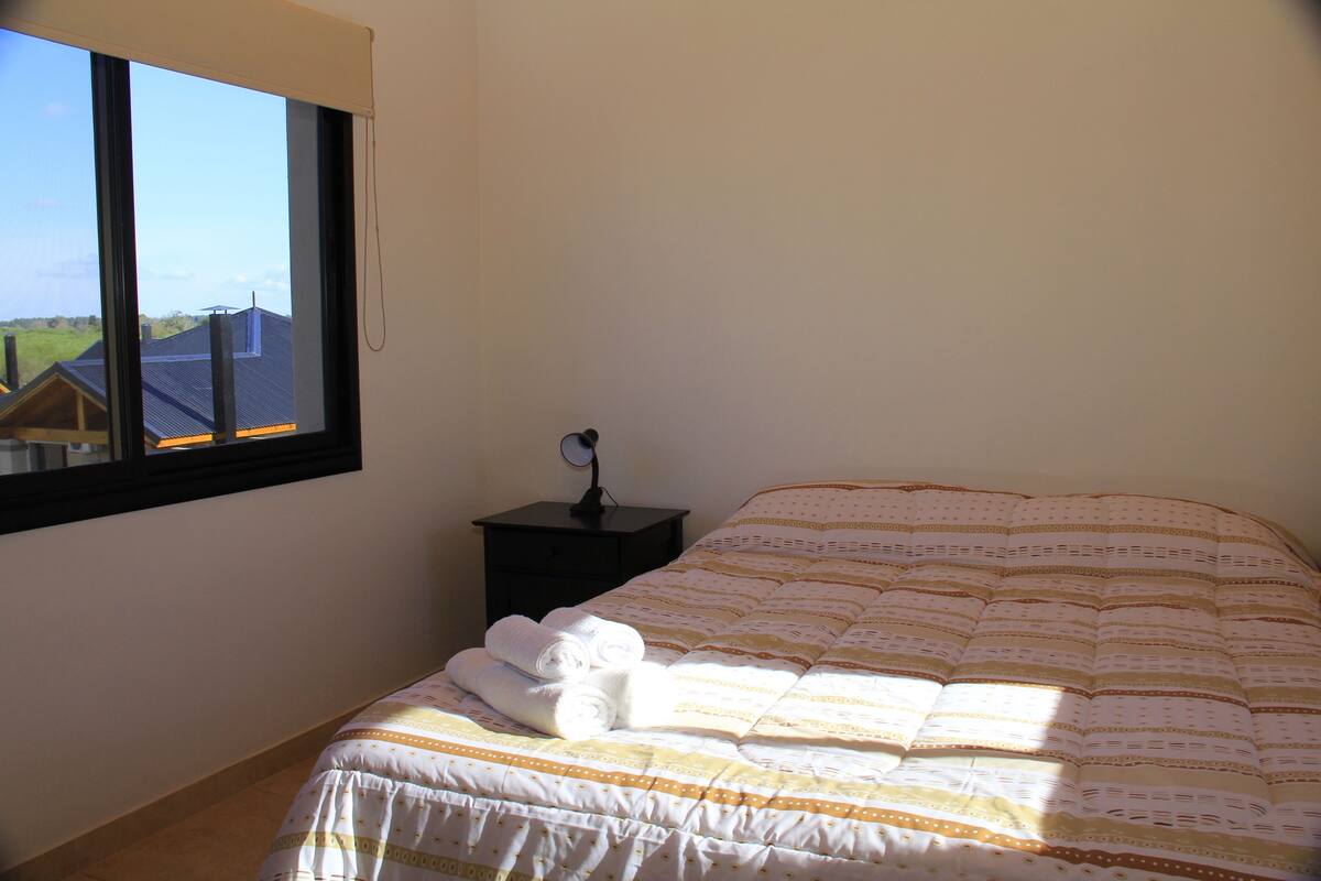 A bedroom with a large window, a bed with a patterned bedspread, a nightstand with a lamp, and a view of a house outside.