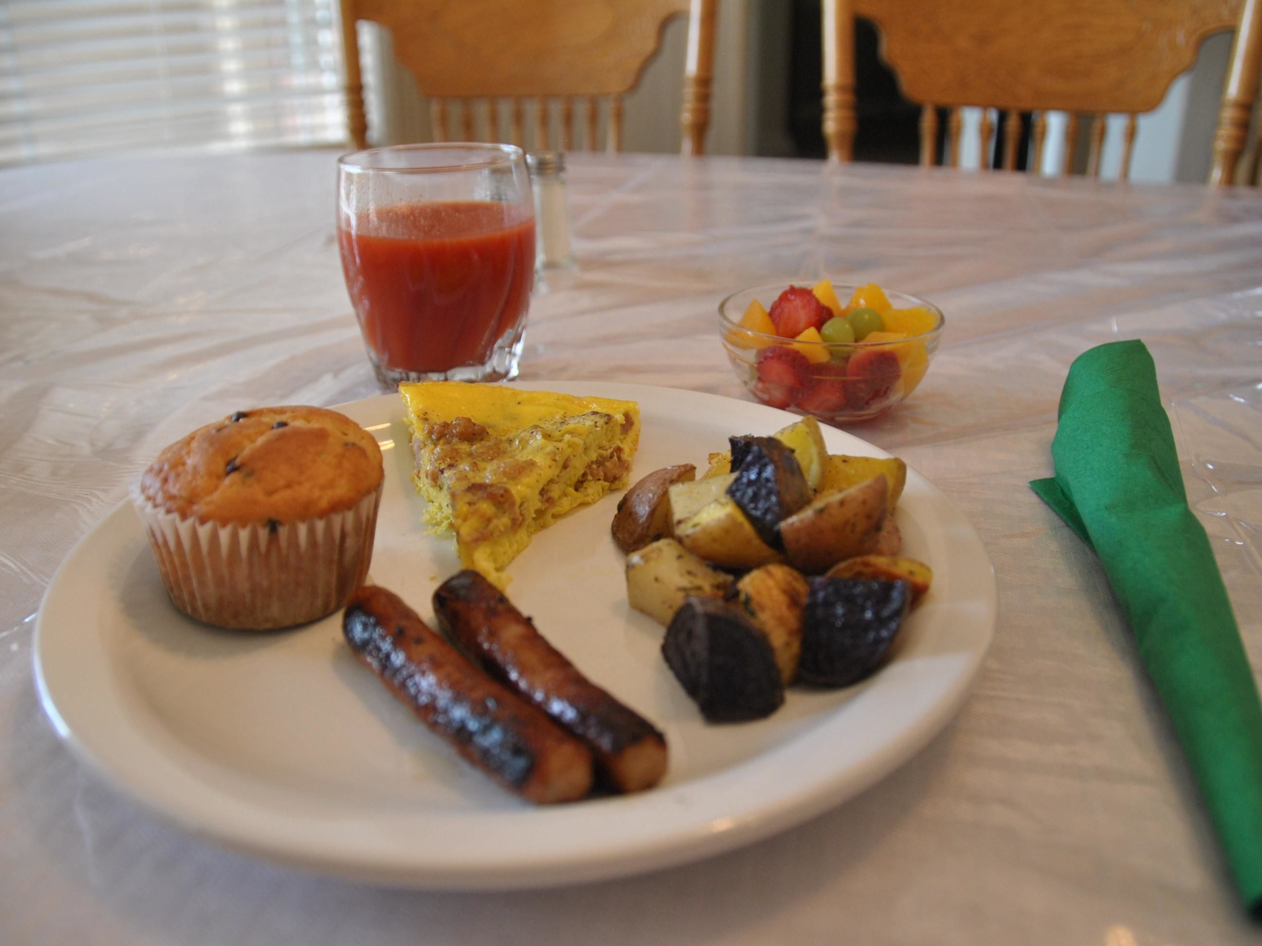 A breakfast plate with a muffin, sausage, omelette, roasted potatoes, and a fruit bowl.